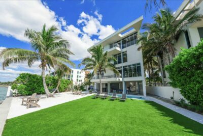 Modern three-story waterfront home in Sarasota with palm trees, artificial turf lawn, a white sand lounge area, and a lap pool along the water's edge.